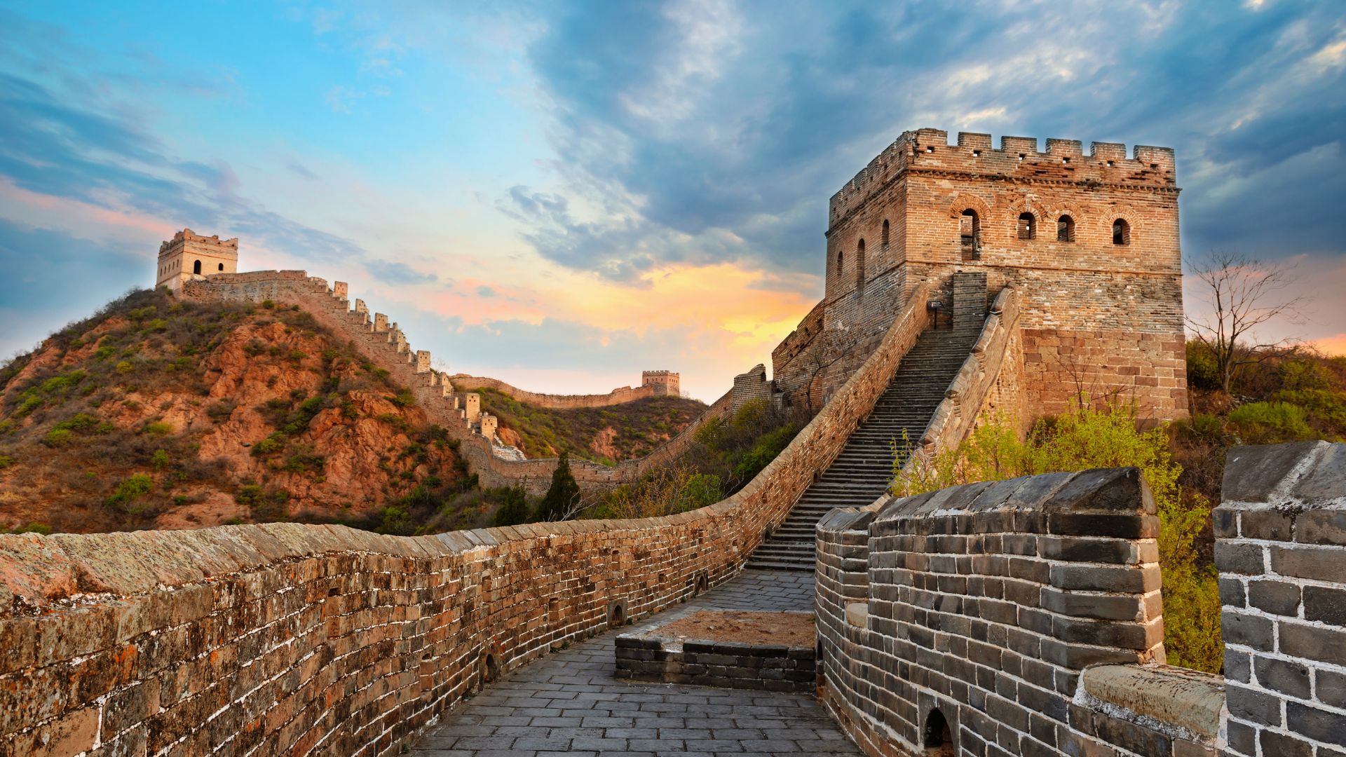 Panoramic view of the Great Wall of China winding across green mountains under a sunset sky.