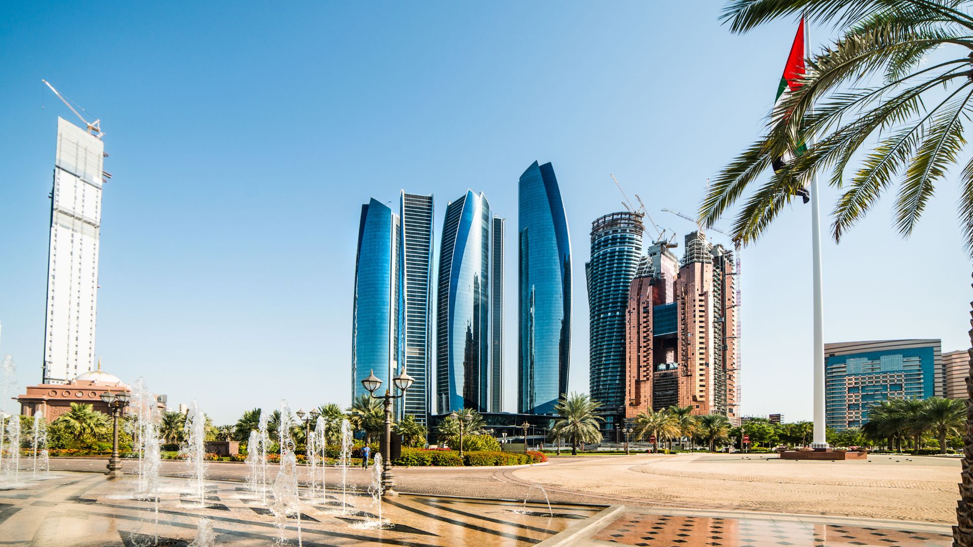 Sunny daytime view of the Etihad Towers skyline in Abu Dhabi, with a plaza fountain spraying in the foreground and the UAE flag flying on the right.