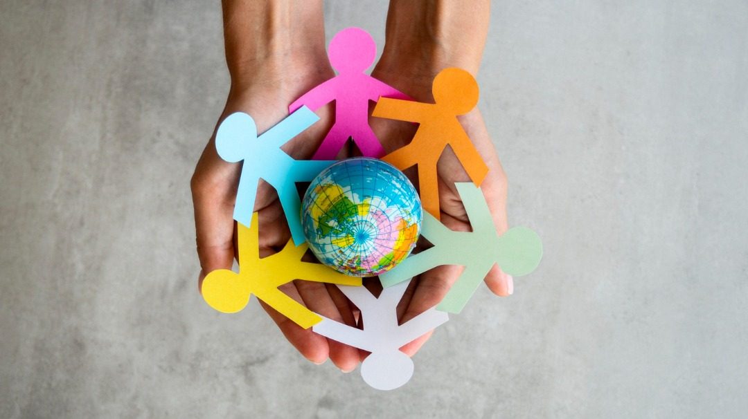 Hands holding a small globe surrounded by colorful paper cut-out figures of people, symbolizing unity, diversity, and global connection.