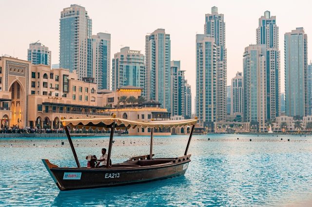 A traditional wooden abra boat sailing on the water with the modern Dubai Marina skyscraper skyline in the background.
