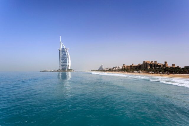 The Burj Al Arab hotel standing on a coastline next to a sandy beach and blue ocean water in Dubai.
