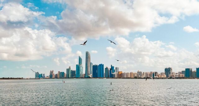Abu Dhabi city skyline with modern skyscrapers viewed across the water under a cloudy blue sky.