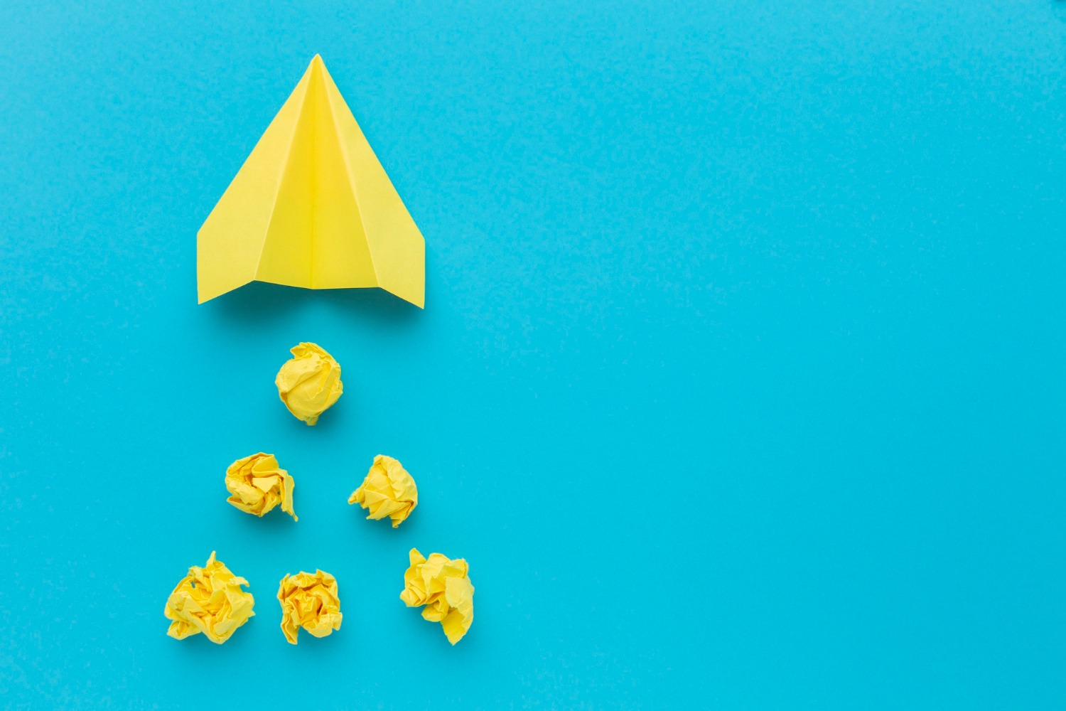 Yellow paper airplane flying above crumpled yellow paper balls on a bright blue background, creating a whimsical scene of paper flight and scattered objects.