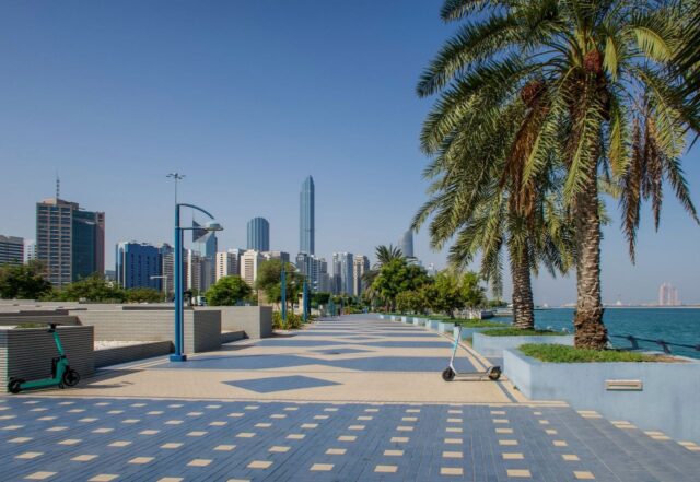Waterfront promenade at the Abu Dhabi Corniche with palm trees, electric scooters, and the city skyline in the background.