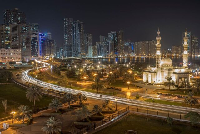 The illuminated Al Noor Mosque in Sharjah at night with light trails from traffic and the city skyline in the background.