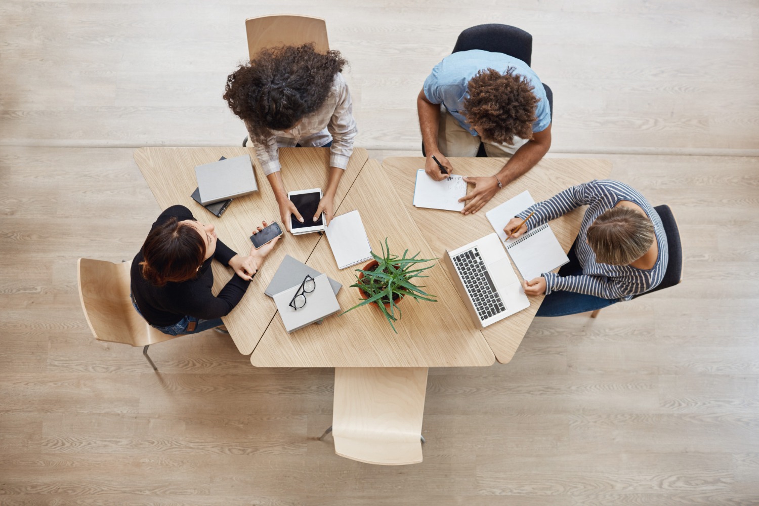 Overhead view of a diverse group of four people working around a wooden table. They are using laptops, tablets, and notebooks, with a small plant in the center of the table. The individuals are engaged in a collaborative discussion or brainstorming session.