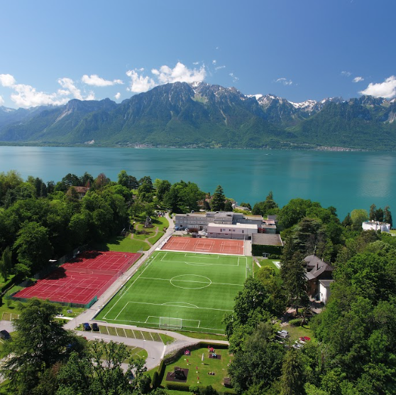 Aerial view of a lakeside international school campus with sports fields, surrounded by greenery and mountains overlooking a turquoise lake on a clear sunny day.