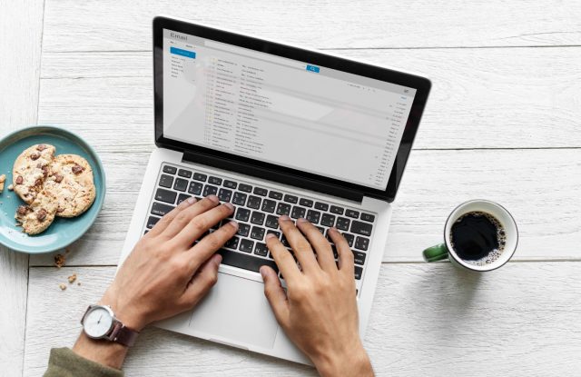 Hands typing on a laptop with an open email inbox on screen, next to a plate of cookies and a cup of coffee on a white wooden desk.