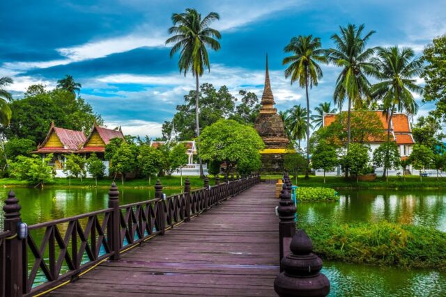 A scenic view of a Thai temple with a traditional spired chedi nestled among tropical palm trees, reflected in a tranquil pond with a dark wooden footbridge leading toward the temple complex.