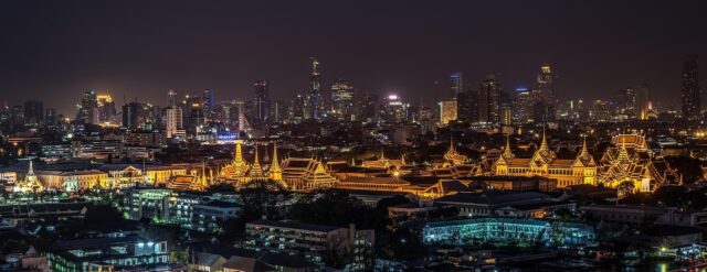 Panoramic night view of the illuminated Grand Palace complex in Bangkok, Thailand, set against the dark city skyline and urban buildings.