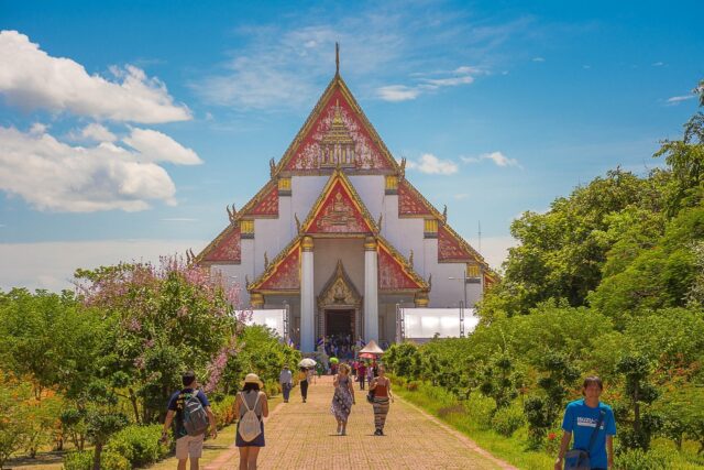 People walking along a brick pathway toward a traditional Thai temple with tiered roofs, surrounded by lush green trees under a bright blue sky with light clouds. (international school job in Thailand)