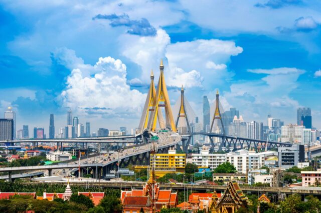 Cityscape of Bangkok showing modern skyscrapers, busy overpass highways, and a grand suspension bridge set against a bright blue sky with light clouds, with traditional Thai temple roofs visible in the foreground.