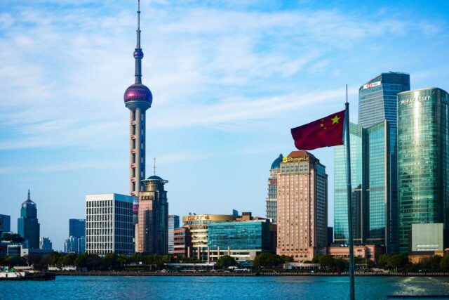 Daytime skyline view of modern Shanghai skyscrapers, including the Oriental Pearl Tower, with the Chinese national flag fluttering in the foreground.