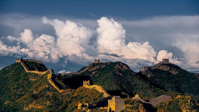 Panoramic view of the Great Wall of China winding across steep mountain ridges under a cloudy blue sky.
