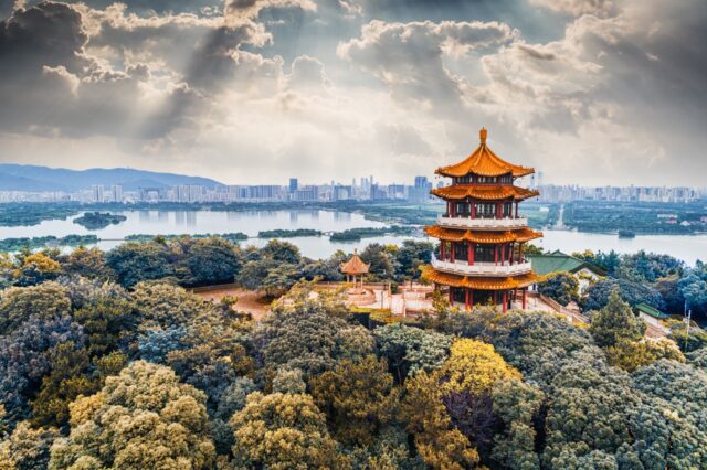 Traditional multi-tiered Chinese pagoda standing amidst autumn foliage overlooking a wide river and modern city skyline under dramatic sunlight.