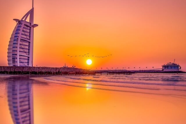 The Burj Al Arab hotel in Dubai at sunset with the sun reflecting on the ocean.
