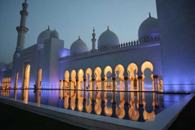 Sheikh Zayed Grand Mosque at night illuminated in purple light with reflections on water.