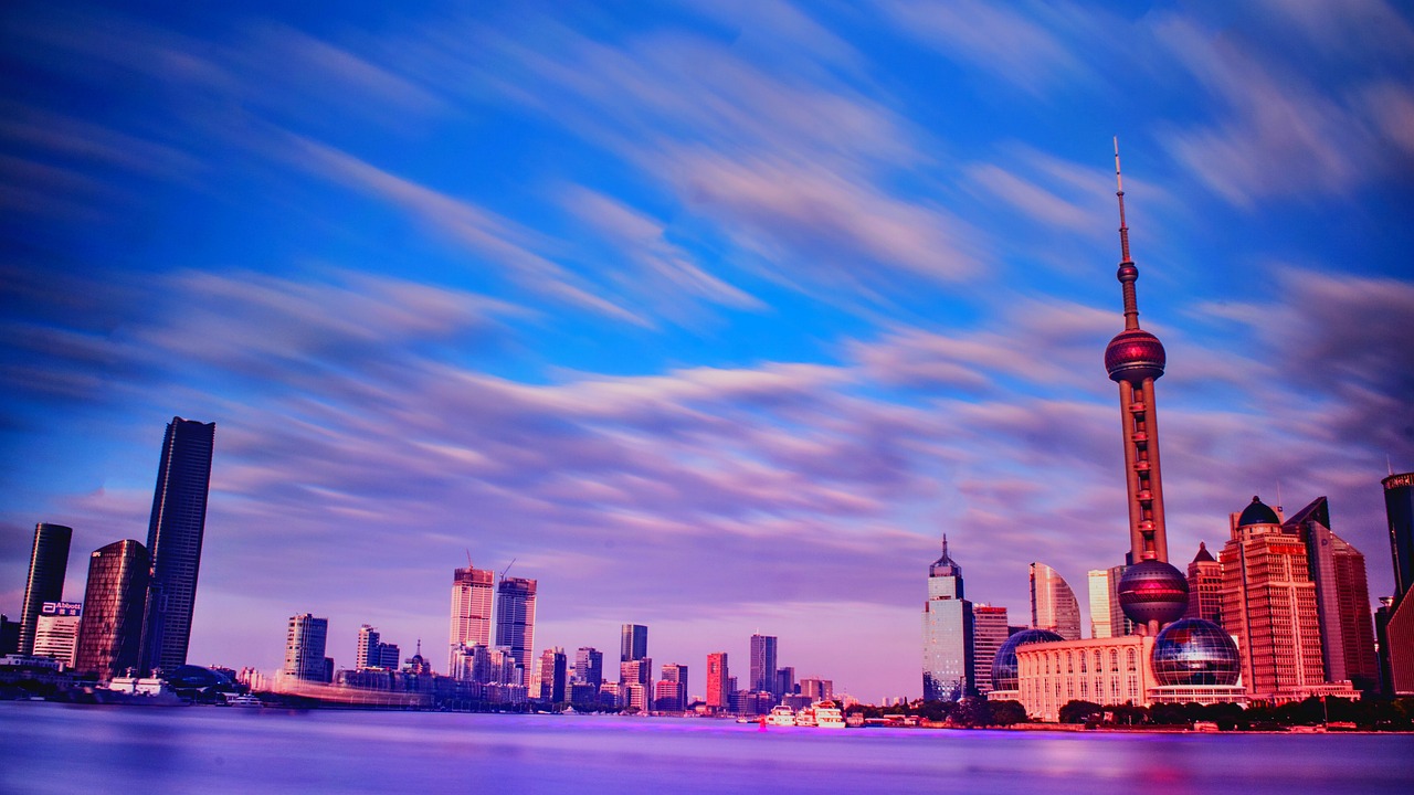 A vibrant twilight view of the Lujiazui financial district in Shanghai across the water, with the city lights casting a colorful glow under streaky, fast-moving blue clouds.