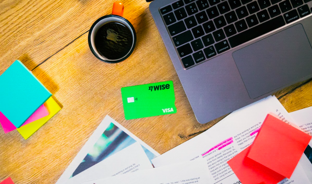 A workspace with a laptop, a green Wise card, colorful sticky notes, coffee mug, and scattered papers on a wooden table.