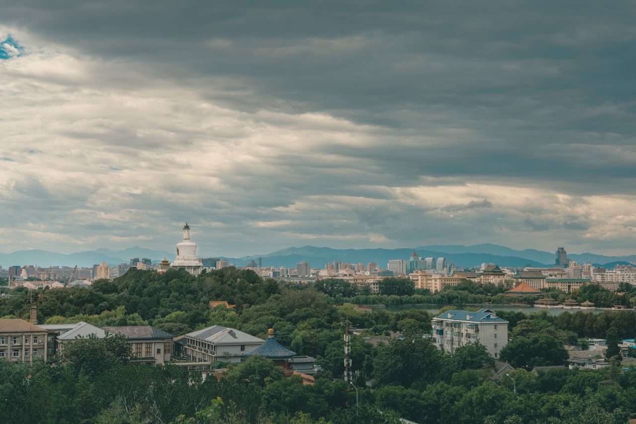 The historic white Tibetan-style stupa of Beihai Park rising above a dense canopy of green trees, contrasting with the modern high-rises of Beijing stretching into the background.