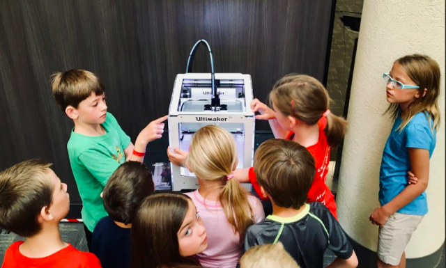 A group of children observing a 3D printer as it creates an object, showcasing their interest in technology and innovation.