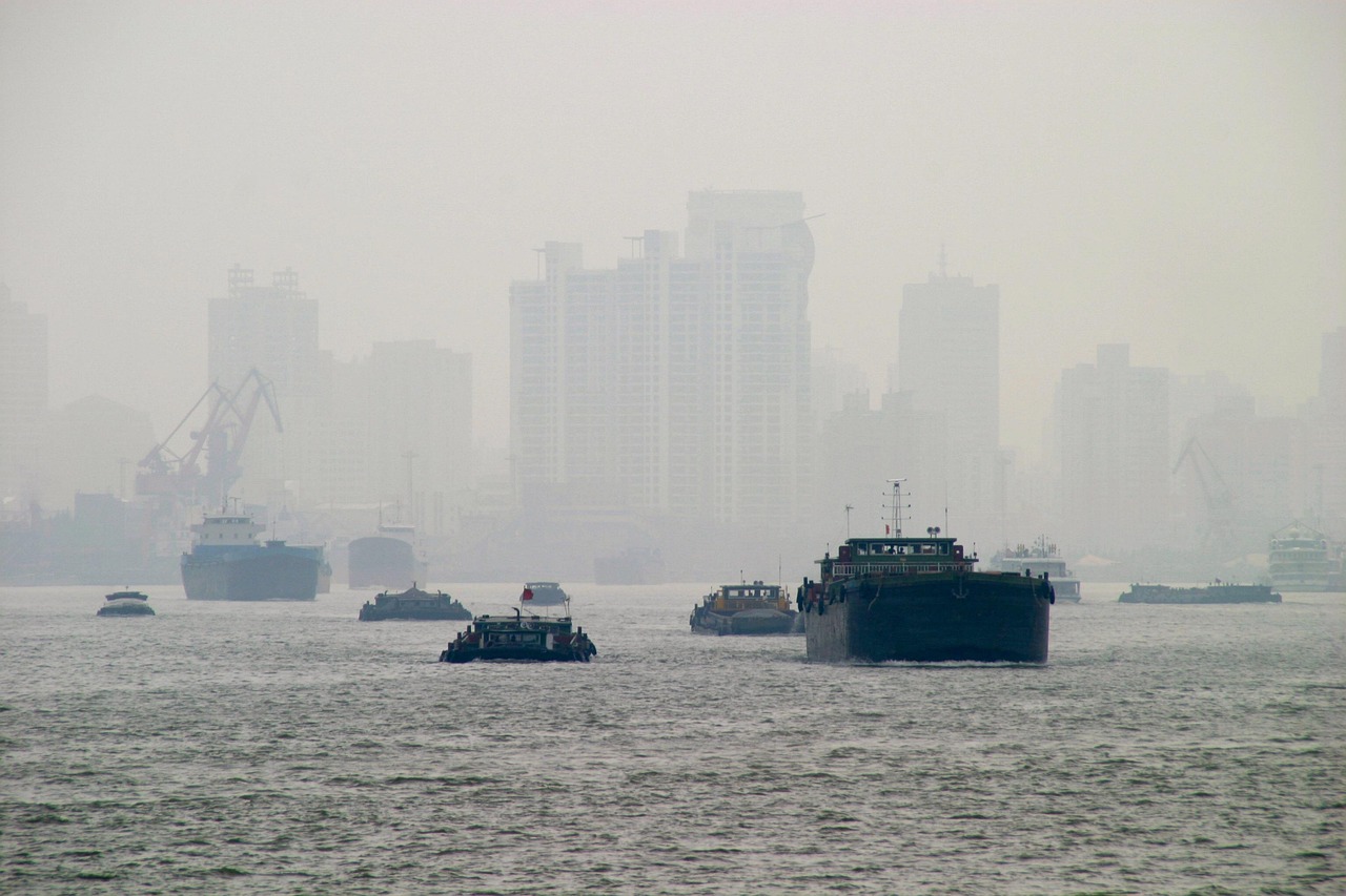 Large industrial boats navigating a river on a heavily smoggy and overcast day, with a cityscape obscured by thick haze in the background.