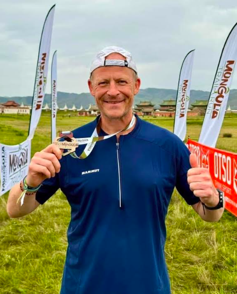 Steve Hill, a passionate teacher, holding a medal and giving thumbs-up at the Mongolia Sunrise to Sunset event, surrounded by event flags.