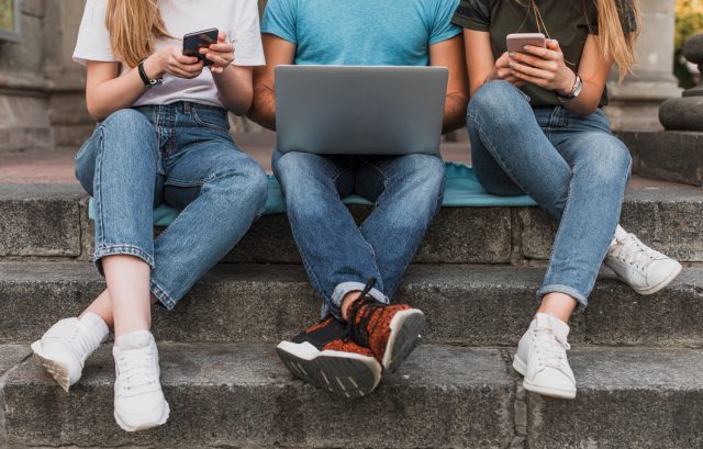 Three individuals seated on steps, each using laptops and cell phones, engaged in conversation and work.