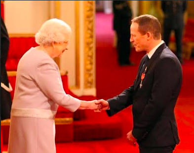 Steve Hill, a dedicated teacher and Deputy Headteacher, pictured with Queen Elizabeth II against a luxurious red carpet and ornate decor.