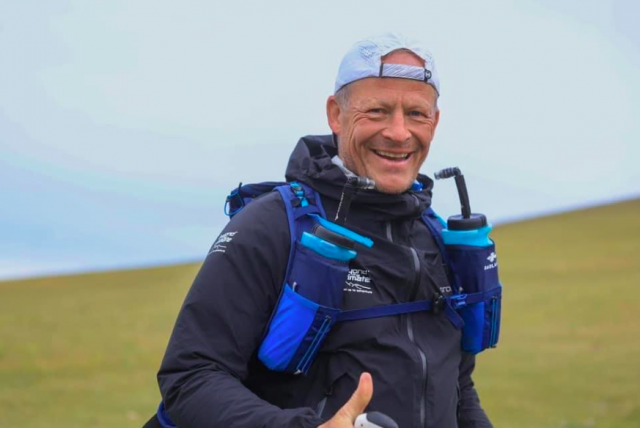Steve Hill, an adventurous educator, hiking in a grassy landscape wearing a black jacket and white cap with a hydration backpack, giving a thumbs-up.