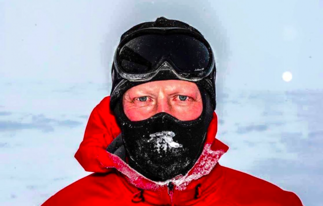Steve Hill, a resilient teacher, braving a snowy landscape in a red jacket and black face mask with goggles, facing a pale sun during a North Pole trek.