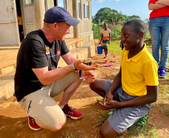 Steve Hill, a compassionate teacher, crouching to engage with a child in a yellow shirt outside a Ugandan school, supported by Team Hill Charitable Trust efforts.