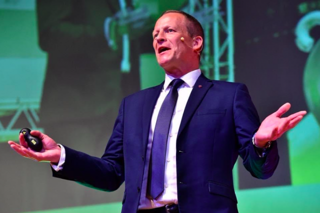 Steve Hill, an inspiring educator and leader, speaking energetically on stage in a blue suit with arms outstretched, holding a remote against a green backdrop.